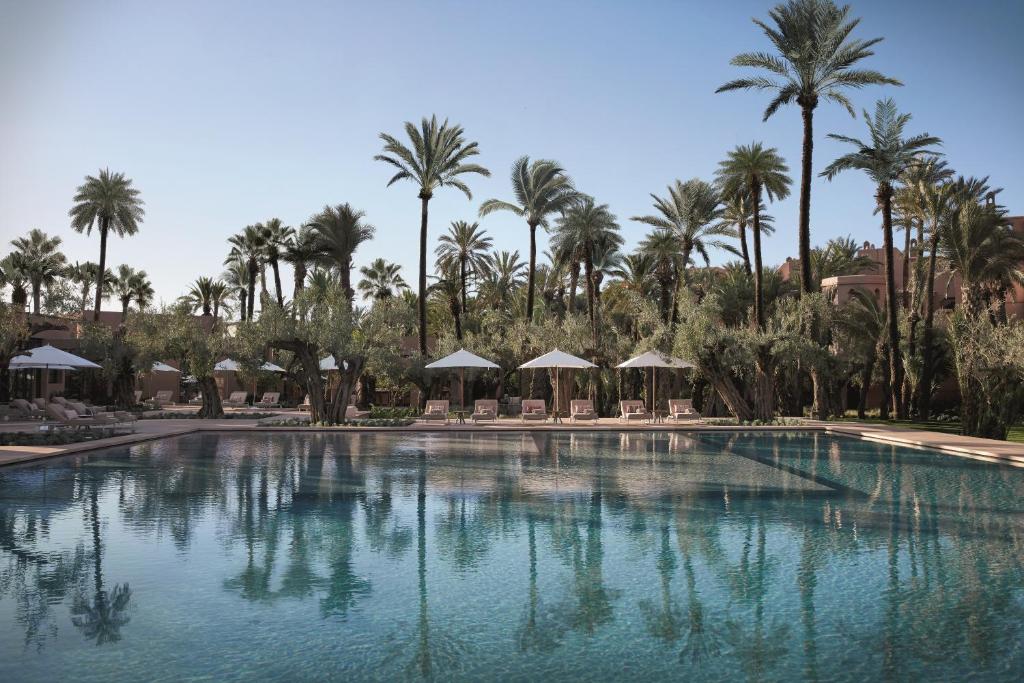 10 Unique Riads in Marrakech with Pool 36 A large, clear swimming pool is surrounded by lounge chairs with white umbrellas and tall palm trees under a clear blue sky, evoking the relaxing, tropical resort vibes of riads in Marrakech with pool.