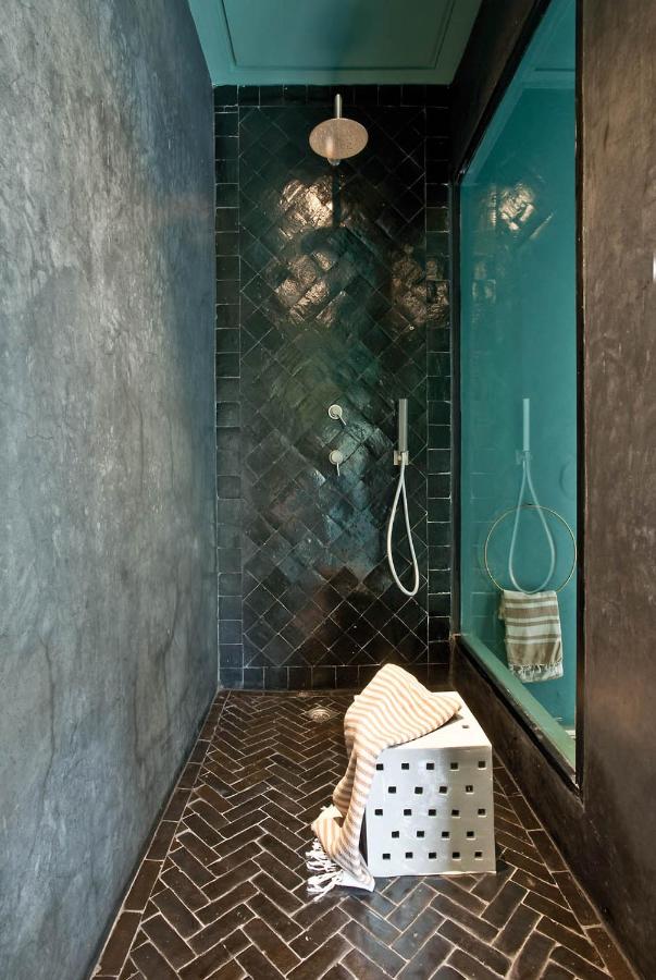10 Unique Riads in Marrakech with Pool 13 A modern shower with glossy black herringbone tiles, a large overhead showerhead, and a built-in niche evokes the elegance of riads in Marrakech with pool. A glass wall separates the area, while a silver cube stool holds a striped towel.