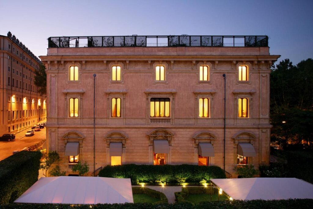 13 UNIQUE Boutique Hotels in Rome 20 A three-story neoclassical building with large lit windows at dusk, surrounded by greenery and white canopies, stands at a street corner—reminiscent of boutique hotels in Rome, Italy.