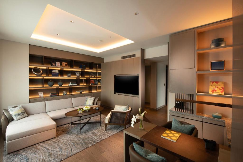 7 Best Hotels in Hiroshima, Japan 19 Modern living room with a beige sectional sofa, a round coffee table, and a wall-mounted TV—echoing the inviting comfort of the best hotels in Hiroshima. Built-in shelves display decor, and warm ambient lighting fills the space, with a dining table in the foreground.