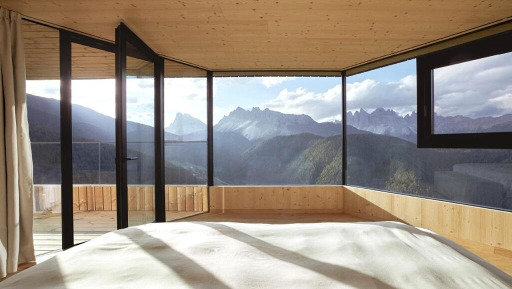 A modern wooden bedroom with large floor-to-ceiling windows offers a panoramic view of distant mountains, green hills, and a partly cloudy sky—an experience reminiscent of the best hotels in the Dolomites. Sunlight softly illuminates the bed and interior space.