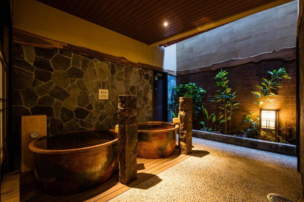 15 Unique Hotels in Tokyo: Coolest hotels 16 A Japanese-style indoor bath area in one of the unique hotels in Tokyo, featuring two large round tubs, stone walls, wooden ceiling, pebbled floor, and a small garden with green plants and a glowing lantern outside. Warm, relaxing atmosphere.