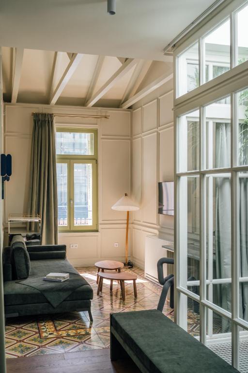 A living room inspired by boutique hotels in Athens, featuring a dark green sectional sofa, geometric patterned floor tiles, and a large window with white mullions and grey curtains.