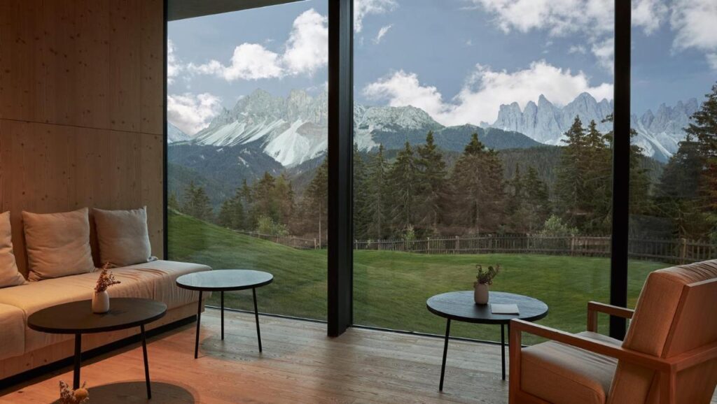 A modern living room with large floor-to-ceiling windows offers a stunning view of pine trees and snow-covered mountains, reminiscent of the best hotels in the Dolomites. Minimalist sofas, chairs, and small round tables complete the serene space.