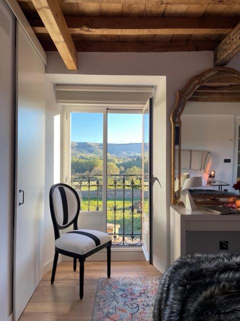 A cozy bedroom with a wooden ceiling, a white chair by a window overlooking green hills, a large mirror on a vanity, and sunlight streaming in—reflecting the charm found in many boutique hotels in Cantabria.