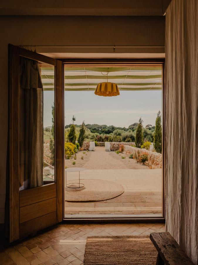 Unique Boutique Hotels in Menorca 19 View from inside a room through open wooden doors to a sunny patio and garden with trees and shrubs, under a partially striped awning—a tranquil scene often found in boutique hotels in Menorca. A yellow pendant light hangs above the doorway.