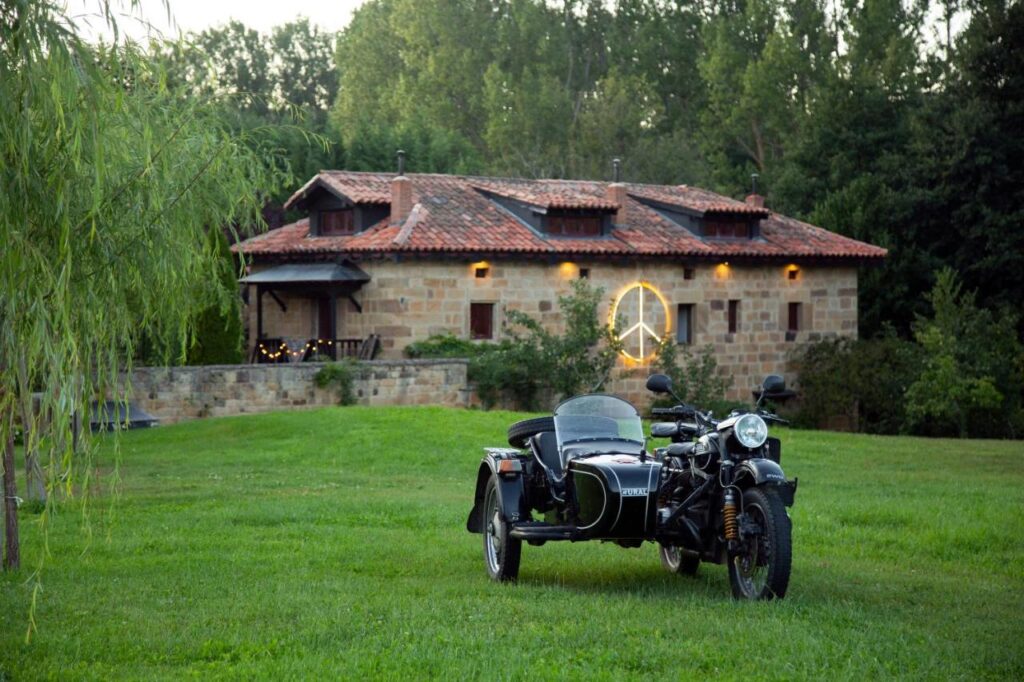 A black motorcycle with a sidecar is parked on grass before a rustic stone house with red roof tiles—similar to charming hotels in Cantabria—and a glowing peace sign on the wall, all surrounded by trees and lush greenery.