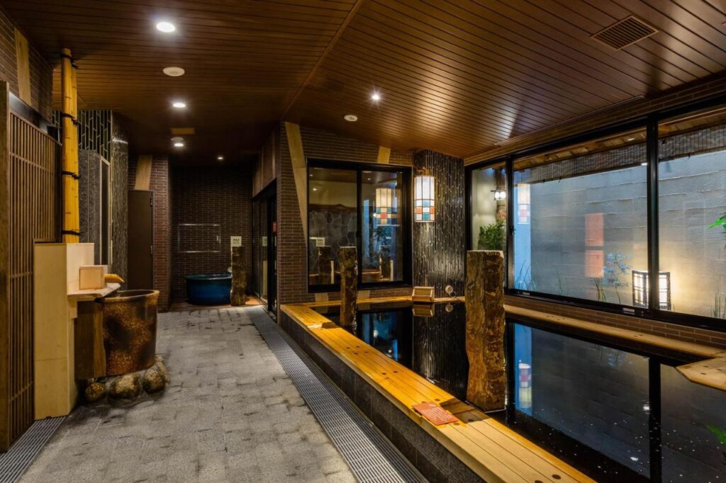15 Unique Hotels in Tokyo: Coolest hotels 15 A serene indoor Japanese onsen at one of the unique hotels in Tokyo, featuring wooden benches, stone flooring, warm lighting, bamboo accents, and a reflecting pool beside large windows to create a relaxing atmosphere.
