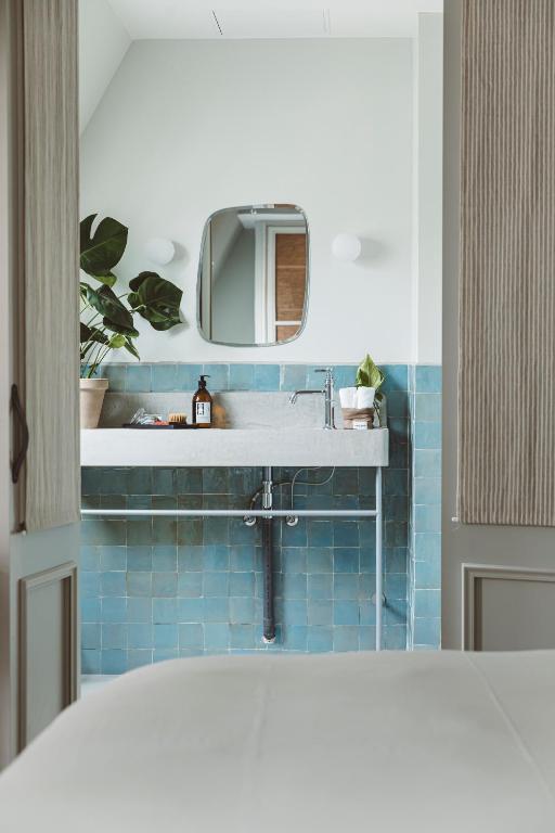 13 Unique Hotels in Paris for a Stylish Stay 30 A modern bathroom in the style of unique hotels in Paris, with blue-green tiled walls, a large white sink, a square mirror, round lights, potted plants, and toiletries on the counter, viewed through an open doorway.