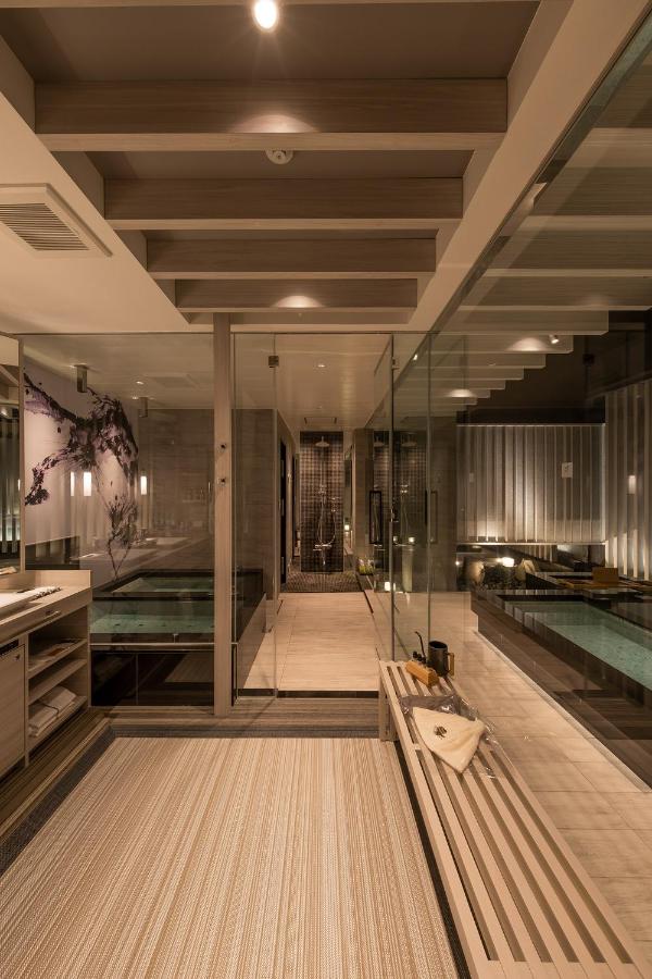 15 Unique Hotels in Tokyo: Coolest hotels 37 Modern, luxurious bathroom inspired by unique hotels in Tokyo, with glass walls, a wooden bench, large bathtub, spacious walk-in shower, warm lighting, and minimalist decor. The floor has a neutral striped rug and shelves with towels.