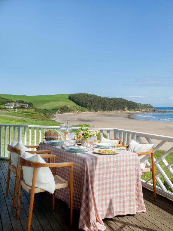 A table covered with a pink gingham cloth sits on a wooden deck overlooking a sandy beach, green hills, and the ocean under a clear blue sky—a scene reminiscent of charming hotels in Cantabria. Four cushioned wooden chairs surround the table.