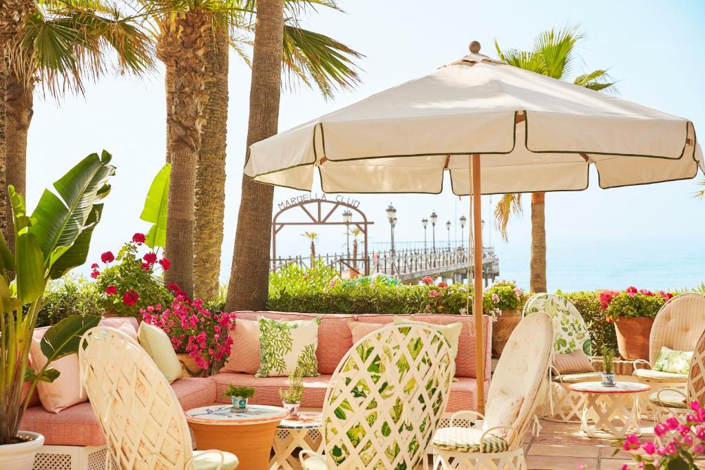 TOP 10 Luxury Hotels in Andalucía 21 Outdoor patio with white wicker chairs, pink cushions, potted plants, and a large umbrella. Overlooking the sea and palm trees, this spot captures the elegance often found at luxury hotels in Andalucía.