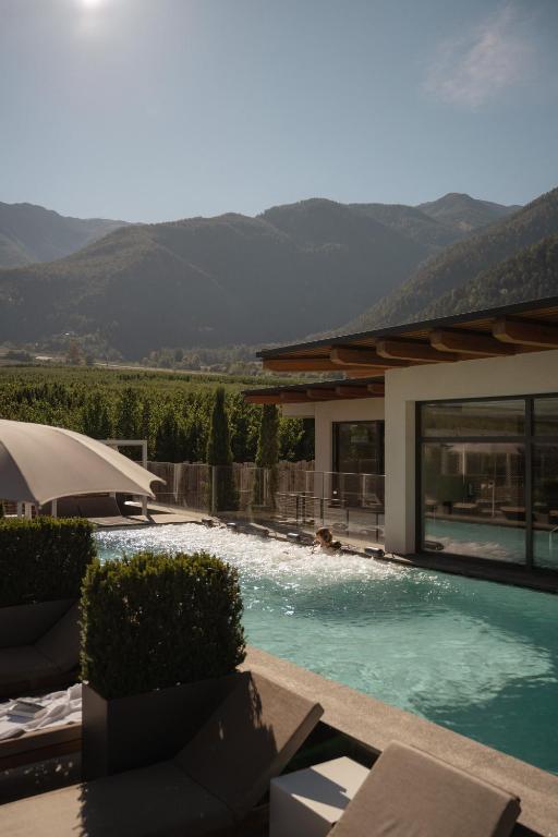 A person swims in a modern outdoor pool beside a building at one of the best hotels in the Dolomites, with lounge chairs and umbrellas nearby. Green hills and mountains create a stunning backdrop under a clear sky.