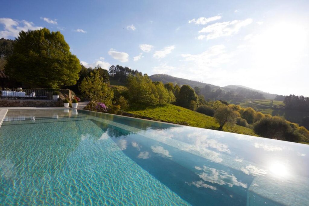 An infinity pool at one of the hotels in Cantabria overlooks a lush green landscape with rolling hills and trees under a bright blue sky dotted with scattered clouds.