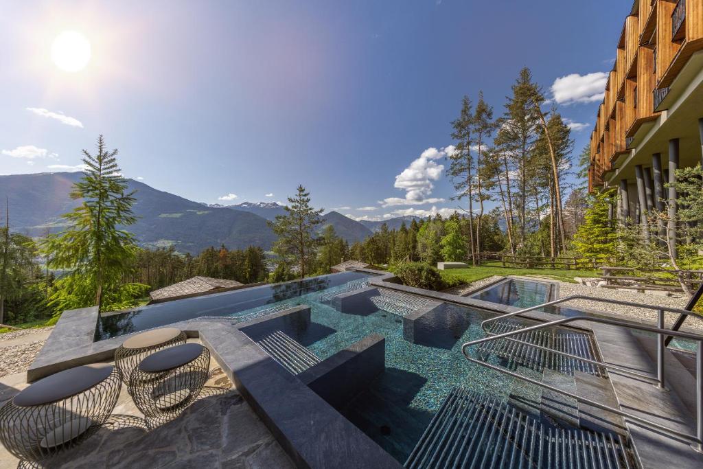 An infinity pool with built-in loungers overlooks forested hills and distant mountains under a bright sunny sky, beside a modern wooden building with outdoor seating—one of the best hotels in the Dolomites for scenic relaxation.