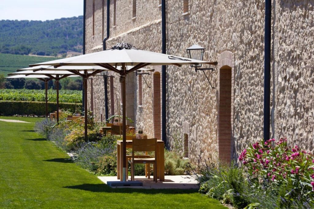 4 Best Hotels in Ribera del Duero, Spain 4 Outdoor patio with wooden tables and chairs under large umbrellas by a stone building, bordered by lush grass, vibrant flowers, and a scenic vineyard—perfect for guests seeking hotels in Ribera del Duero.