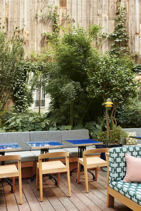 13 Unique Hotels in Paris for a Stylish Stay 19 Outdoor patio with wooden floor, modern wooden chairs, blue tables, patterned bench seating, and lush green plants climbing a tall wooden wall—an inviting spot often found at unique hotels in Paris.