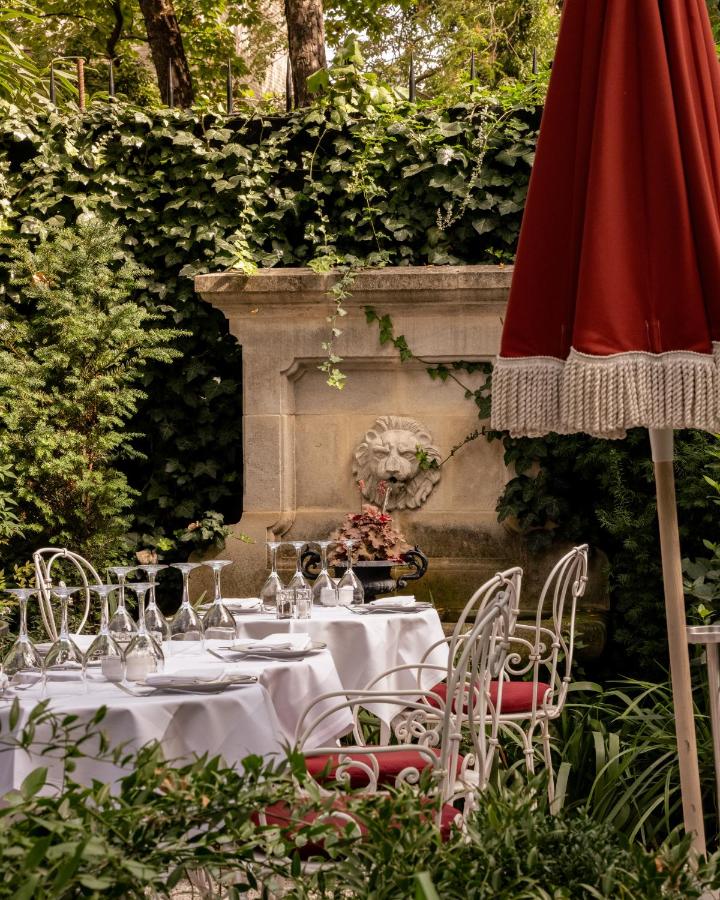 13 Unique Hotels in Paris for a Stylish Stay 10 An elegant outdoor dining area at one of the unique hotels in Paris, featuring white tablecloths, ornate white chairs with red cushions, a red umbrella, lush greenery, and an ivy-covered stone wall with a lion head fountain.