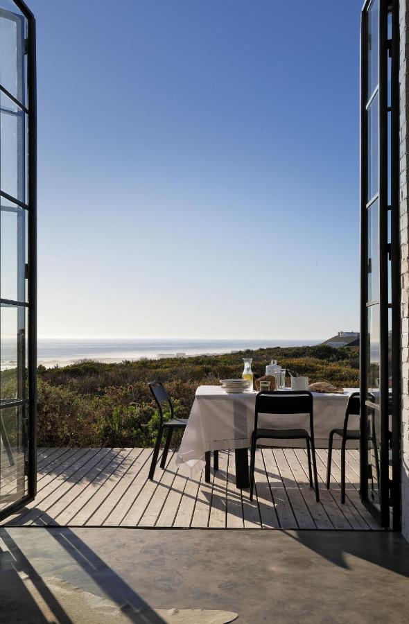 Unique Places to Stay in Cape Town 23 A set table with black chairs sits on a wooden deck outside open glass doors, overlooking green dunes and a calm ocean—one of the unique places to stay in Cape Town under a clear blue sky.