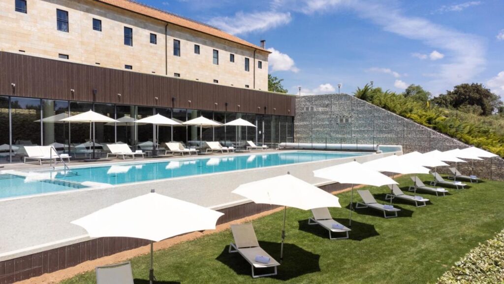 4 Best Hotels in Ribera del Duero, Spain 21 Outdoor pool with sun loungers and white umbrellas on a grassy area, next to a modern building with large windows and stone walls—perfect for relaxing at one of the top hotels in Ribera del Duero under a blue sky with scattered clouds.
