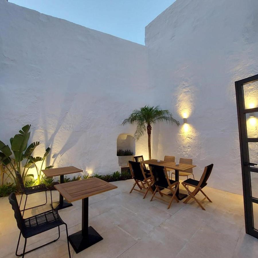 Unique Boutique Hotels in Menorca 32 Outdoor patio with white stucco walls, two wooden tables with chairs, a black bench, potted green plants, and warm wall lights—echoing the serene charm often found in boutique hotels in Menorca—illuminating the area at dusk.