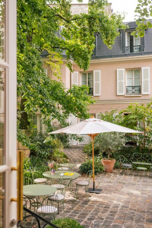 13 Unique Hotels in Paris for a Stylish Stay 43 A charming courtyard at one of the unique hotels in Paris, featuring cobblestone flooring, green metal tables and chairs, a large white patio umbrella, lush greenery, and a peach-colored building with white shutters in the background.