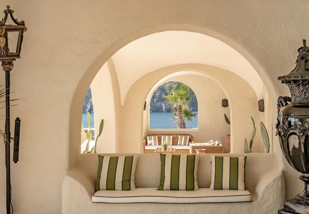 7 Best Hotels in the Aeolian Islands 21 A white stucco alcove with a cushioned bench and green-striped pillows leads to arched openings, revealing an outdoor seating area, palm trees, and a view of the sea—reminiscent of charming hotels in the Aeolian Islands.