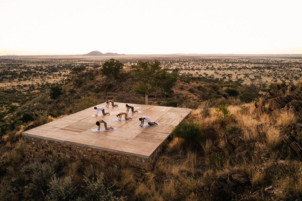 Unique Safari Lodges in Namibia: Wildlife and Adventure 3 A group practices yoga on a wooden platform surrounded by dry grass and sparse trees at one of the safari lodges in Namibia, overlooking a vast savanna landscape under a clear sky.