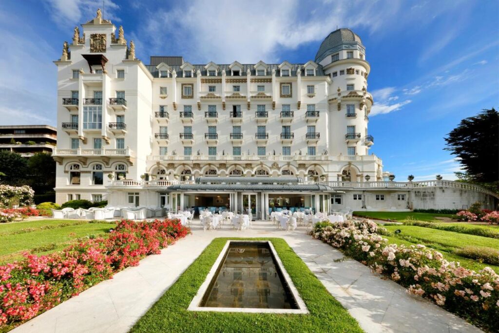 A grand white hotel with ornate balconies and a turret stands behind a lush garden filled with pink and white flowers, manicured hedges, and a narrow reflecting pool—one of the most elegant hotels in Cantabria under a bright blue sky.
