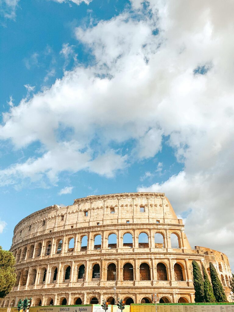 13 UNIQUE Boutique Hotels in Rome 3 The Colosseum in Rome, Italy, stands under a partly cloudy blue sky. The ancient stone amphitheater is partially illuminated by sunlight, with trees and a construction barrier in the foreground—just steps from charming boutique hotels in Rome.