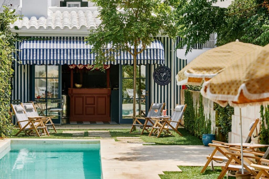 Unique Boutique Hotels in Menorca 6 A backyard scene with a turquoise swimming pool, wooden lounge chairs, striped blue and white awnings, sun umbrellas, and lush greenery evokes the relaxed charm of boutique hotels in Menorca in front of a white house.