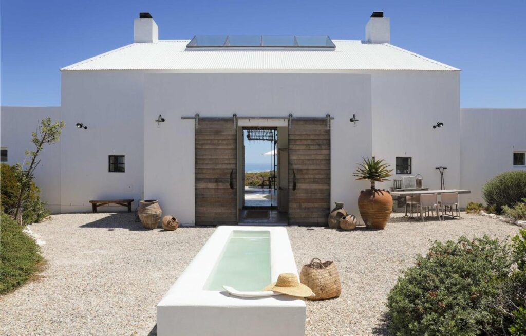 Unique Places to Stay in Cape Town 24 A modern white house with a metal roof, central wooden double doors, and a narrow reflecting pool in front—one of the unique places to stay in Cape Town. Outdoor seating, pottery, and plants decorate the gravel courtyard under a clear blue sky.
