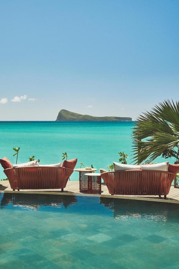 Most Unique Adults Only Hotels in Mauritius, Indian Ocean 4 Two red lounge chairs and a small table sit by a clear blue pool, overlooking the turquoise ocean and a distant island under a sunny sky. A tropical palm frond completes this serene scene at one of the finest adults only hotels in Mauritius.