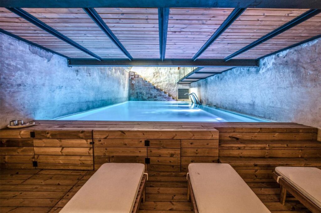 Unique Boutique Hotels in Menorca 29 A modern indoor pool with blue water, surrounded by concrete walls and a wooden ceiling, resembles the serene elegance found in boutique hotels in Menorca. Wooden lounge chairs face the pool, while light enters from stairs at the far end.