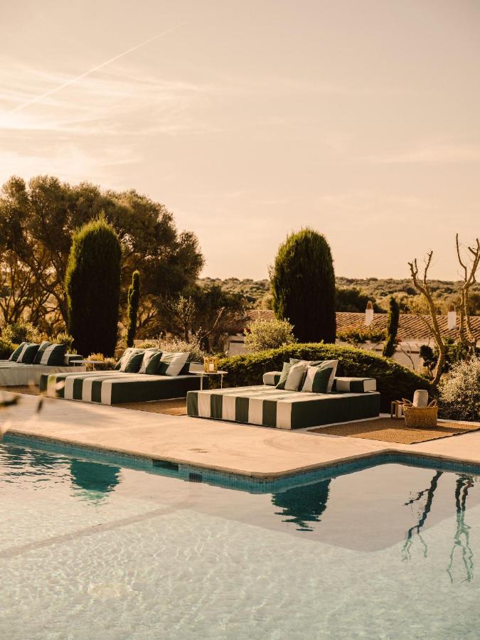 Unique Boutique Hotels in Menorca 13 Lounge chairs with striped cushions sit beside a serene outdoor pool at one of the boutique hotels in Menorca, surrounded by manicured greenery and tall trees under a warm, golden sky. The setting is calm and inviting.