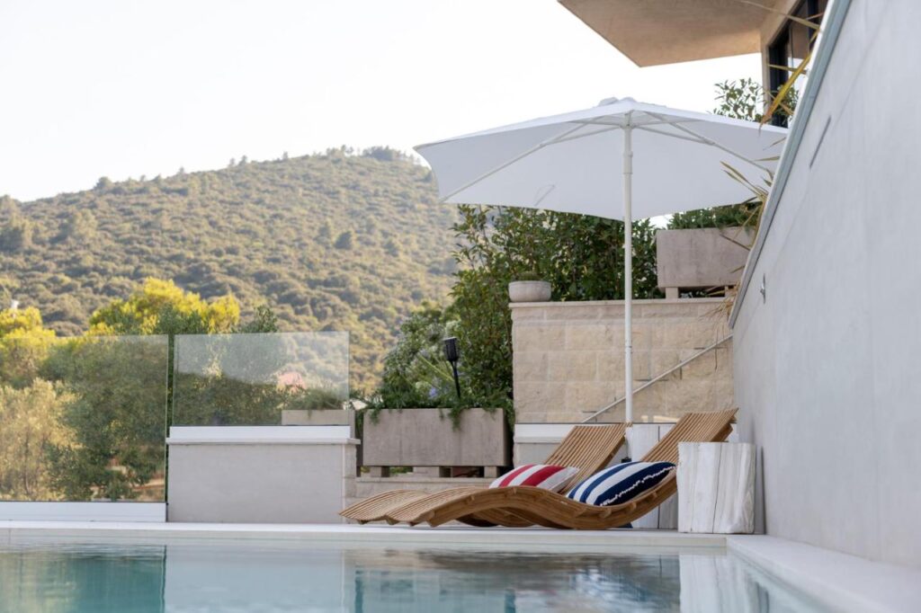 Boutique Hotels in Korčula Island 2 Two wooden lounge chairs with striped cushions sit by a pool at one of the boutique hotels in Korcula, shaded by a white umbrella. The scene overlooks green hills and trees, with a stone wall and glass railing under a clear sky.