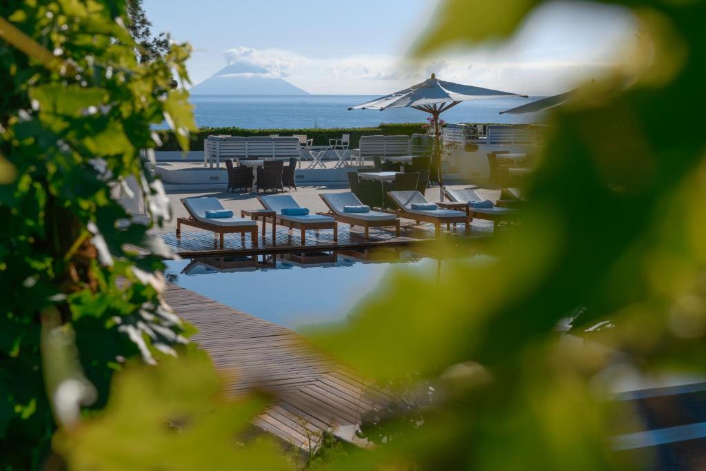7 Best Hotels in the Aeolian Islands 3 View through green leaves of a sunlit pool area with lounge chairs and umbrellas, overlooking the sea and a distant island—classic scenery from some of the best hotels in the Aeolian Islands under a clear blue sky.