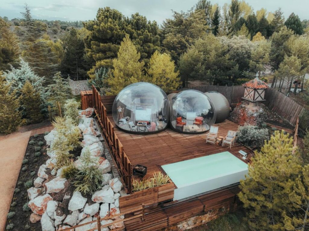 Unique Hotels in Soria: Northern Castile 4 A wooden deck in a forest clearing features two transparent dome tents with lounge chairs and a small rectangular pool, offering a unique escape among trees and rocks—an enchanting alternative to hotels in Soria.