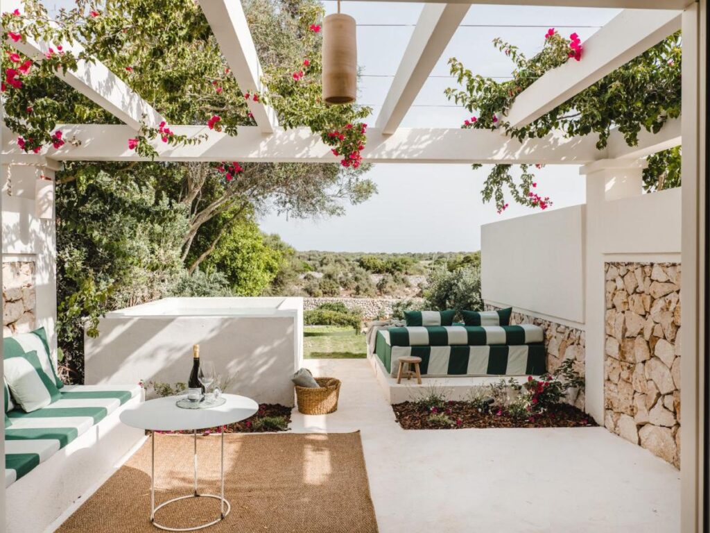 Unique Boutique Hotels in Menorca 11 A stylish outdoor patio at one of the boutique hotels in Menorca features green-and-white striped seating, a round table with wine, stone walls, and flowering vines under a white pergola, overlooking lush greenery and countryside.