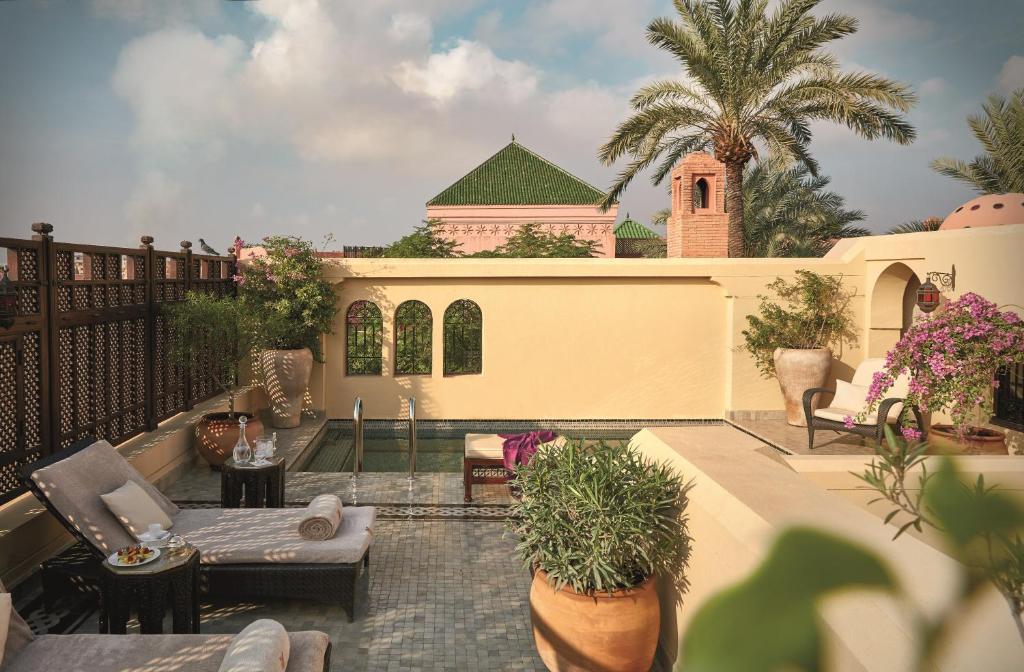 10 Unique Riads in Marrakech with Pool 35 A luxurious outdoor terrace inspired by riads in Marrakech with pool, featuring lounge chairs, potted plants, and flowering shrubs set against palm trees and green-tiled roofs under a partly cloudy sky.