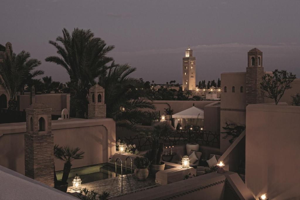 10 Unique Riads in Marrakech with Pool 37 A tranquil rooftop view at dusk shows traditional Moroccan architecture, palm trees, glowing lanterns, and a tall illuminated minaret, capturing the enchanting atmosphere of riads in Marrakech with pool under a dusky sky.