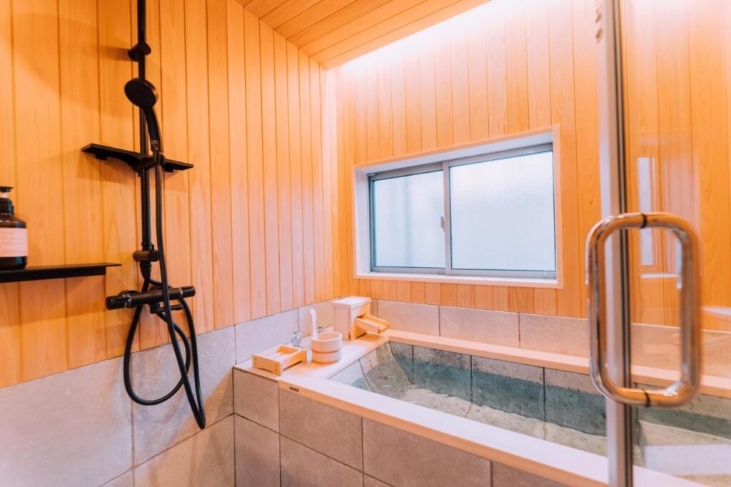 15 Unique Hotels in Tokyo: Coolest hotels 57 A modern Japanese-style bathroom like those found in unique hotels in Tokyo, featuring light wood panel walls, a deep soaking tub, a handheld shower, and a glass door. The minimalist space is brightened by a frosted window letting in natural light.