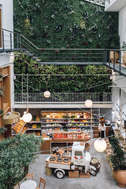 A market stall with fresh fruits, vegetables, and baskets is set indoors beneath a large vertical garden wall, reminiscent of boutique hotels in Athens. A person stands behind the stall, with globed lights above and a small white cart in front.
