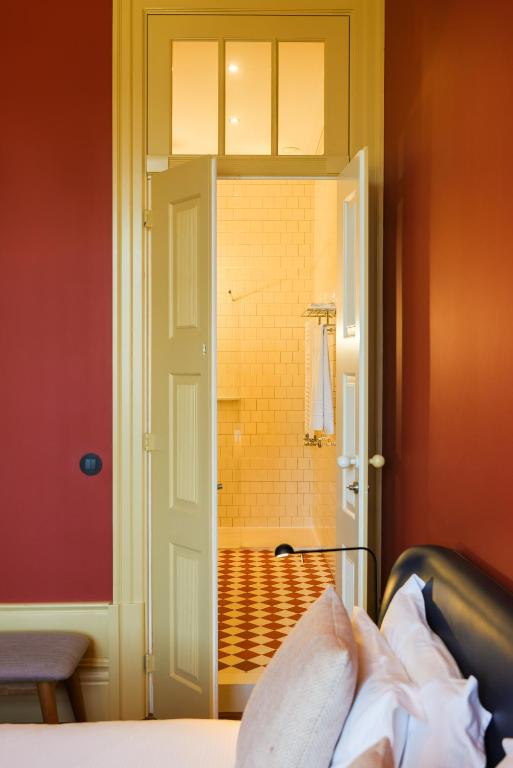 Top 12 Boutique Hotels in Porto, Portugal 30 A cozy room with dark red walls and a bed in the foreground, styled like the finest boutique hotels in Porto. An open cream door leads to a bright bathroom with white tiled walls, a red-and-white checkered floor, and towels neatly hung by the shower.