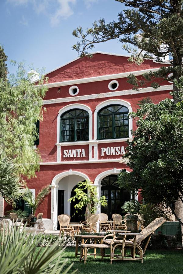 Unique Boutique Hotels in Menorca 27 A red, Mediterranean-style building with arched windows and white trim, labeled Santa Ponsa. Reminiscent of boutique hotels in Menorca, wicker chairs and tables are set on a lush green lawn amid tall trees and plants.