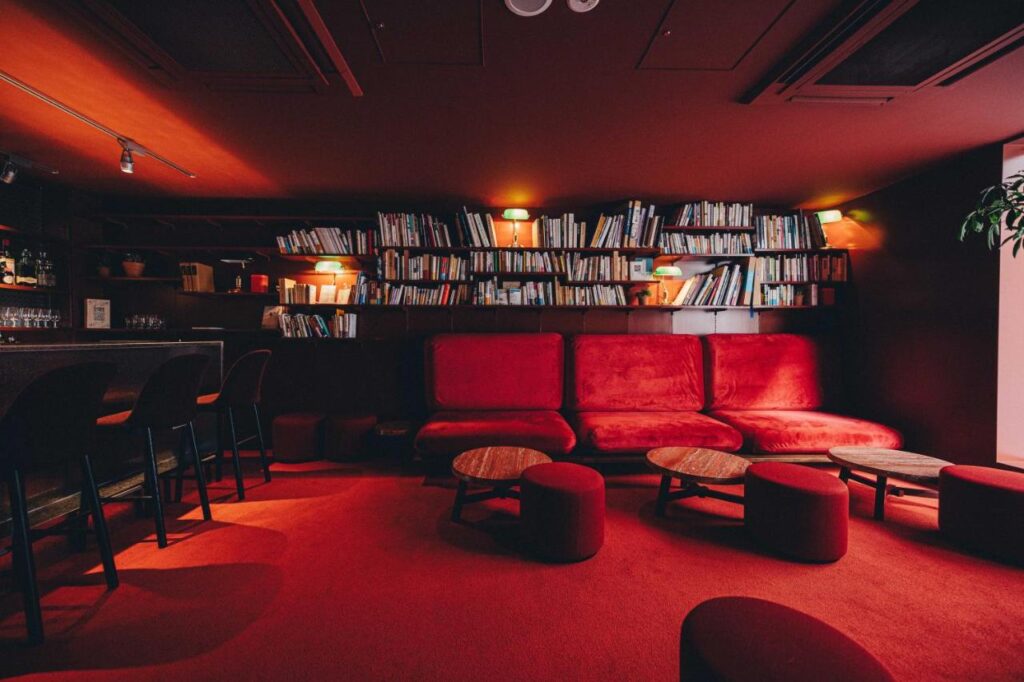 15 Unique Hotels in Tokyo: Coolest hotels 11 A cozy lounge in one of the unique hotels in Tokyo, featuring red carpet and velvet sofas, round wooden tables, dark walls lined with bookshelves, ambient lighting, and a stylish bar area with stools on the left.