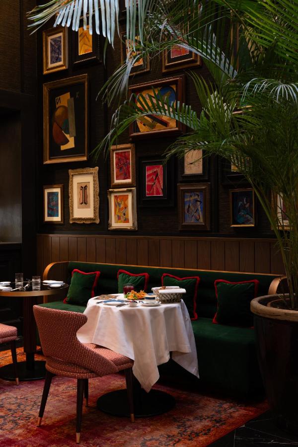 13 UNIQUE Boutique Hotels in Rome 5 A stylish restaurant corner, reminiscent of boutique hotels in Rome, Italy, features a round table for two with pink chairs, a green cushioned bench, potted palm, and colorful gallery wall. Warm, moody lighting enhances the cozy atmosphere.