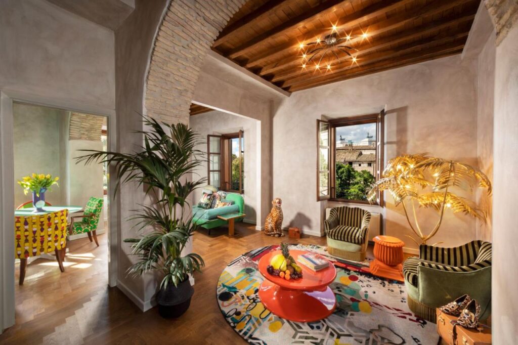13 UNIQUE Boutique Hotels in Rome 36 A vibrant, eclectic living room reminiscent of boutique hotels in Rome, featuring a round colorful rug, green velvet chairs, a gold palm lamp, and sunlit windows. The adjacent dining area boasts a yellow polka dot tablecloth and floral centerpiece.