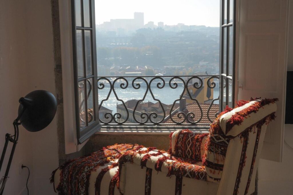 Top 12 Boutique Hotels in Porto, Portugal 45 A cozy armchair with a colorful woven blanket sits by a window, overlooking rooftops, a river, and a hazy cityscape—reminiscent of views from boutique hotels in Porto. Sunlight streams in as a black lamp stands nearby, brightening the inviting space.