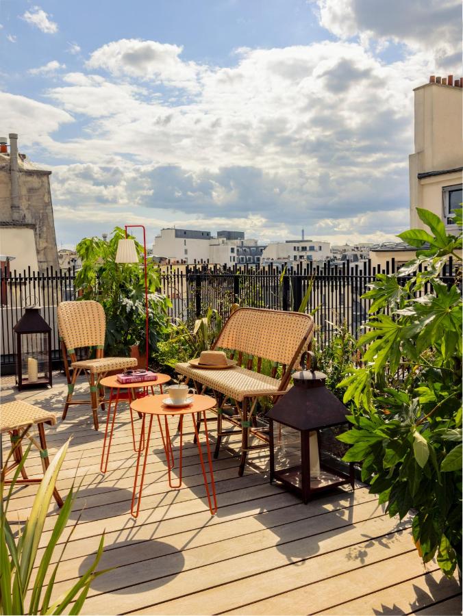 13 Unique Hotels in Paris for a Stylish Stay 8 A sunny rooftop terrace at one of the unique hotels in Paris features wicker chairs, a small sofa, red tables, potted plants, and city buildings in the background under a partly cloudy sky.